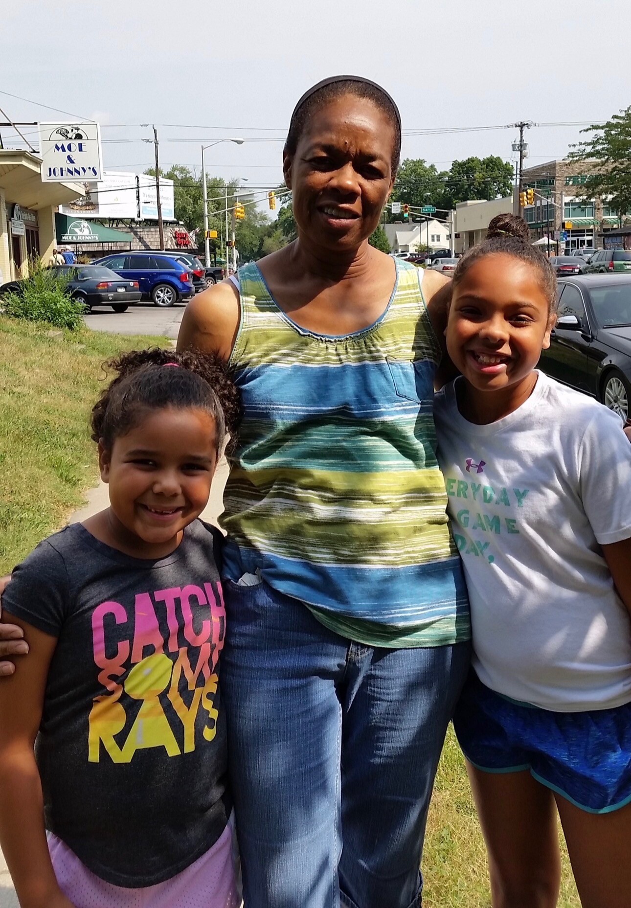 Three people pose for a photo outdoors—an adult woman in the center with two smiling girls on either side, on a sunny street with cars and storefronts in the background.