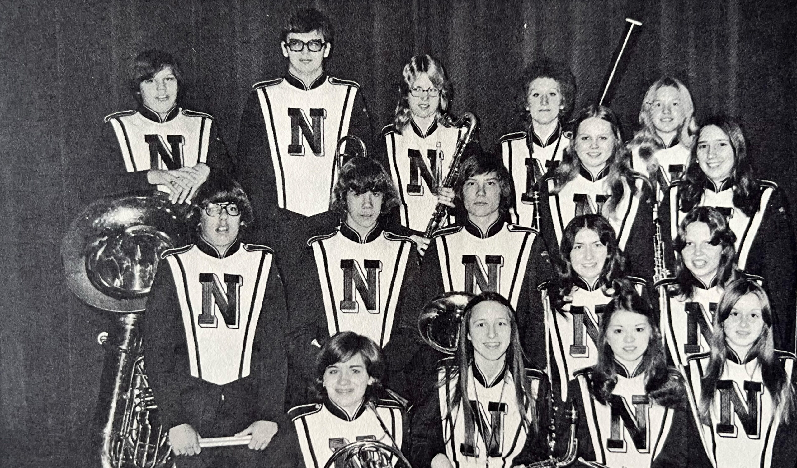 Group of marching band members in matching uniforms with large N on their vests, posing for a class photo on stage with instruments nearby