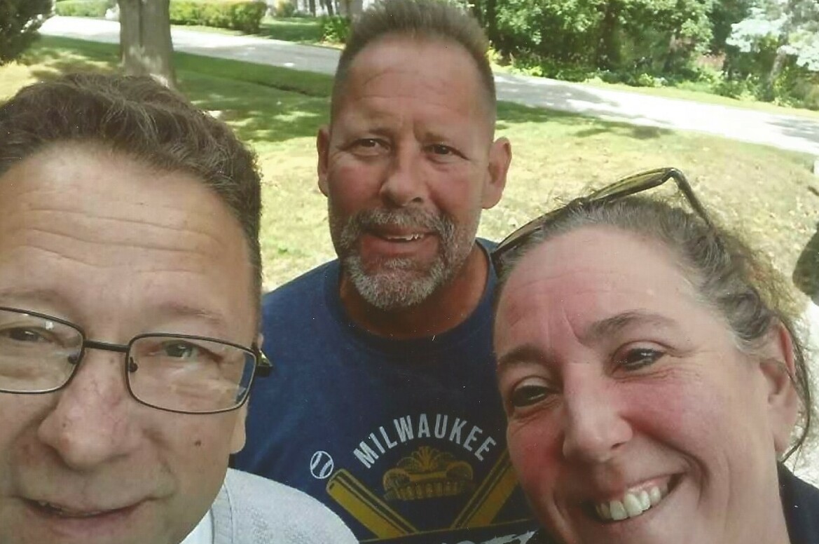 Three adults pose for a sunny outdoor selfie, smiling at the camera with a park and trees in the background in daytime in a casual setting.