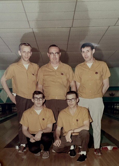 Five men in matching orange bowling shirts pose for a group photo in a bowling alley, with three standing in the back row and two kneeling in front.