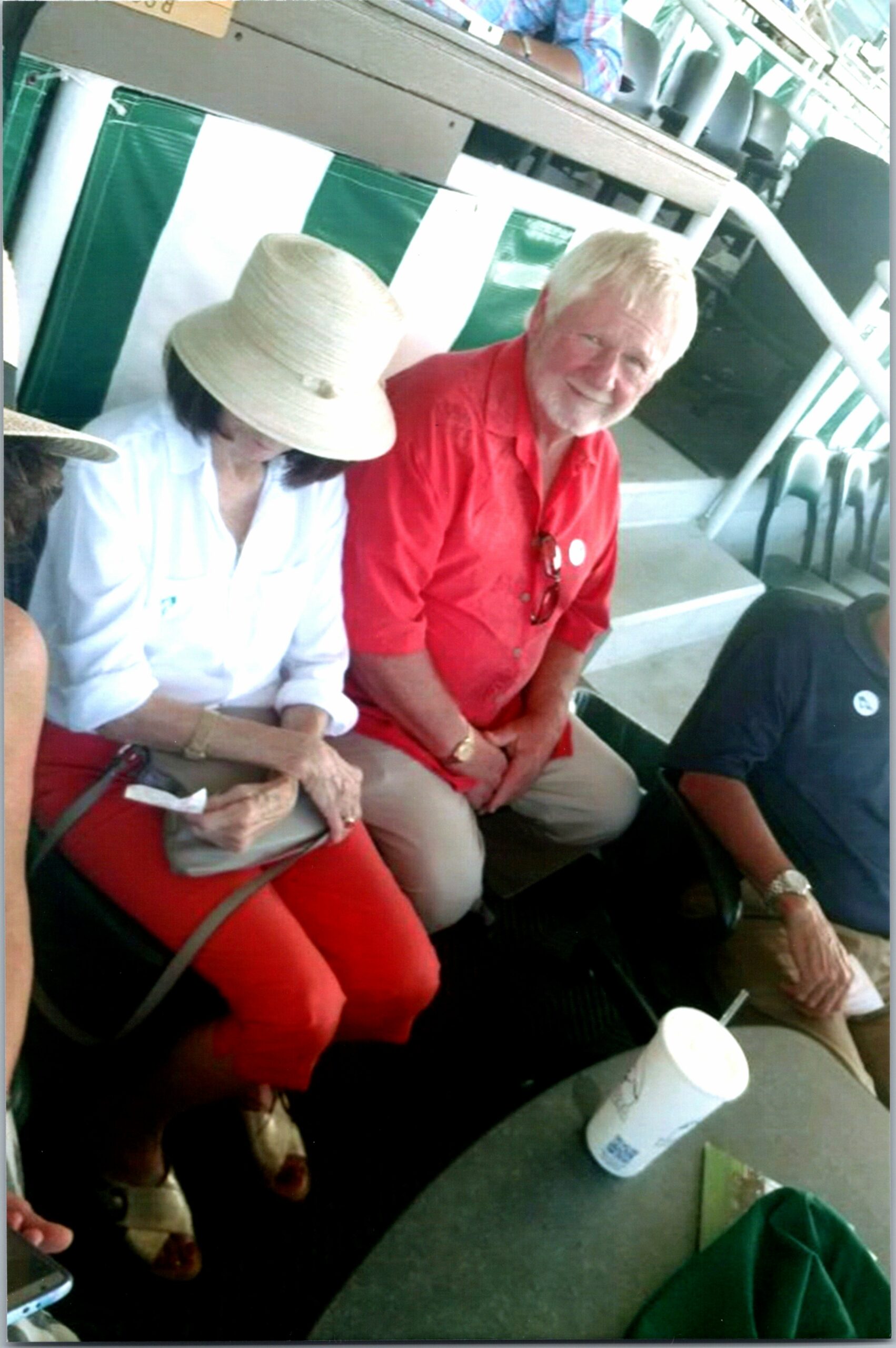 Two older adults sit side by side at an outdoor event, the woman in a white blouse and wide hat and the man in a red shirt smiling nearby.