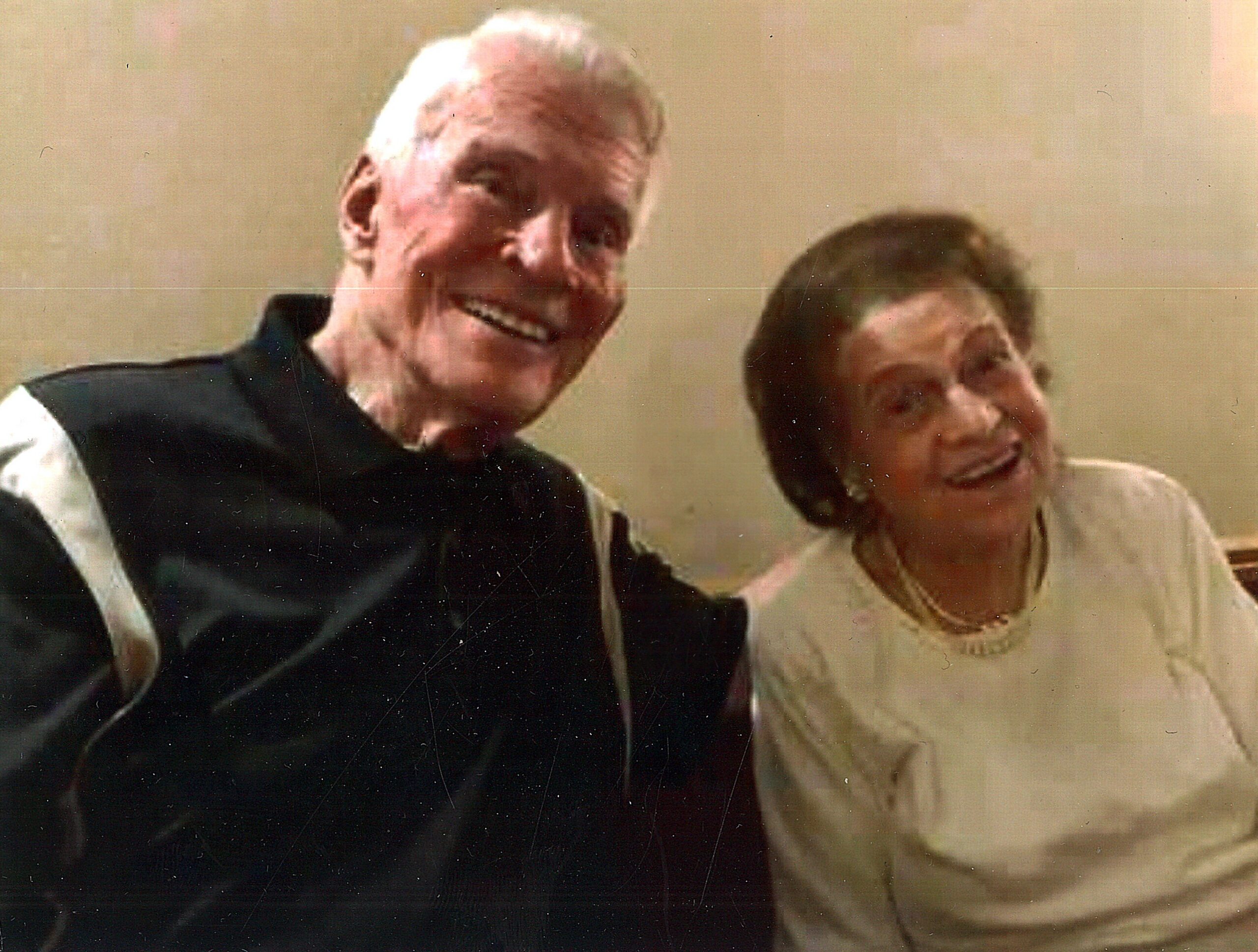 Two smiling older adults posing for a photo, a man in a dark vest beside a woman in a light dress.