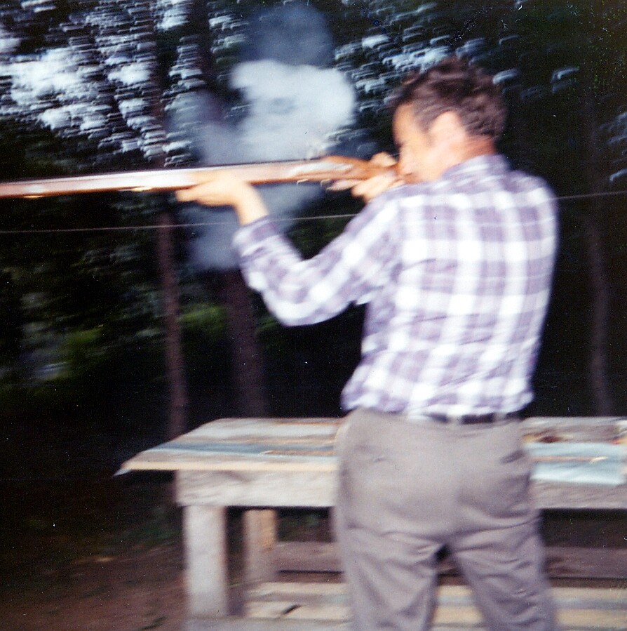 Man in a plaid shirt fires a rifle from shoulder at an outdoor shooting bench, smoke near the muzzle.