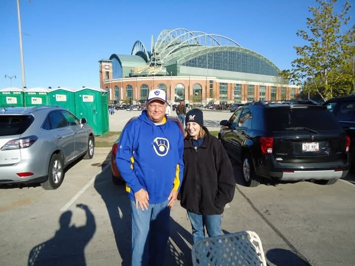 Two people pose in a parking lot in front of a large brick stadium with a curved glass roof; portable toilets visible on the left.