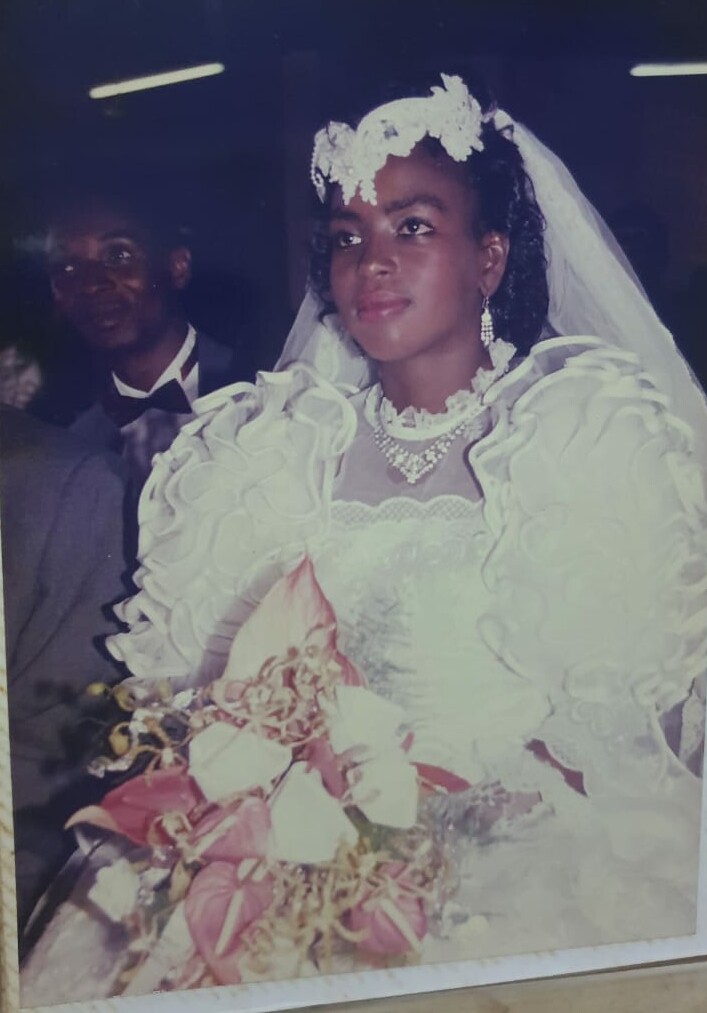 Bride in a white wedding gown with ruffled sleeves and veil, wearing a pearl necklace, holding a pink bouquet.