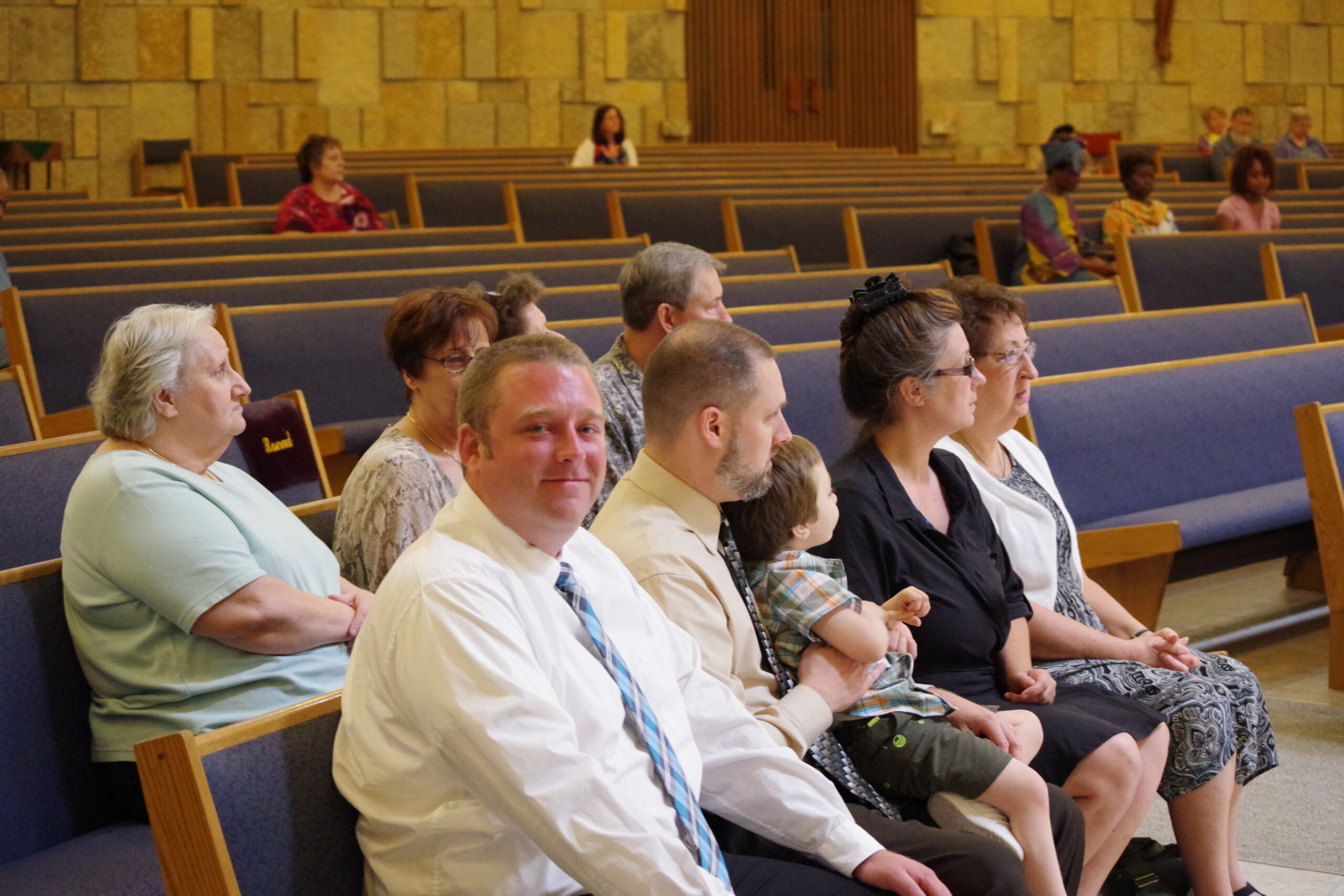 Several adults sit in blue-cushioned church pews, facing forward, in a stone-walled sanctuary.