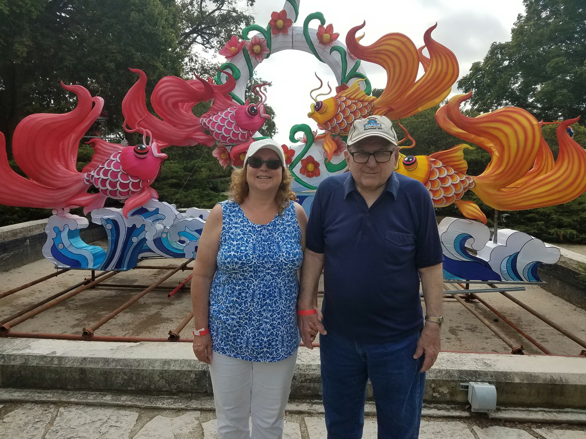 Smiling woman in a blue patterned sleeveless top and a man in a navy shirt pose in front of bright pink and gold dragon boat sculptures at an outdoor display.
