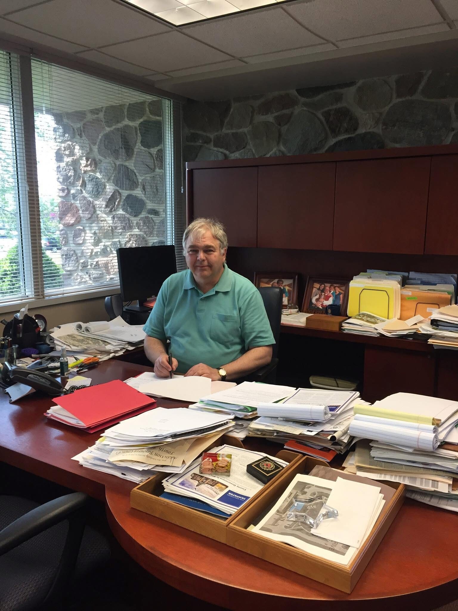 Older man in a teal polo sits at a cluttered office desk, writing with a pen.