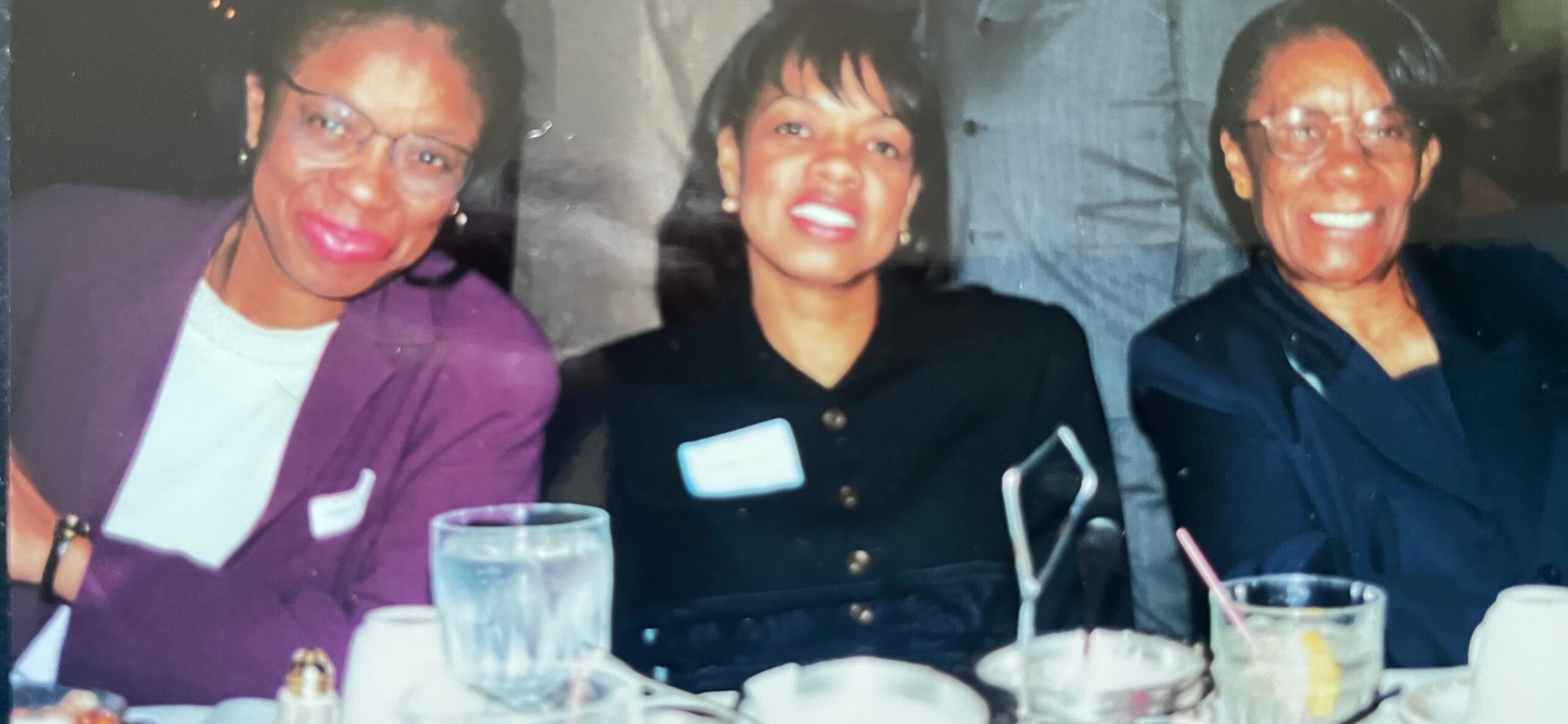Three women smiling at a dinner table with drinks, posing for a photo; left in purple jacket, middle in black, right in dark blue top.