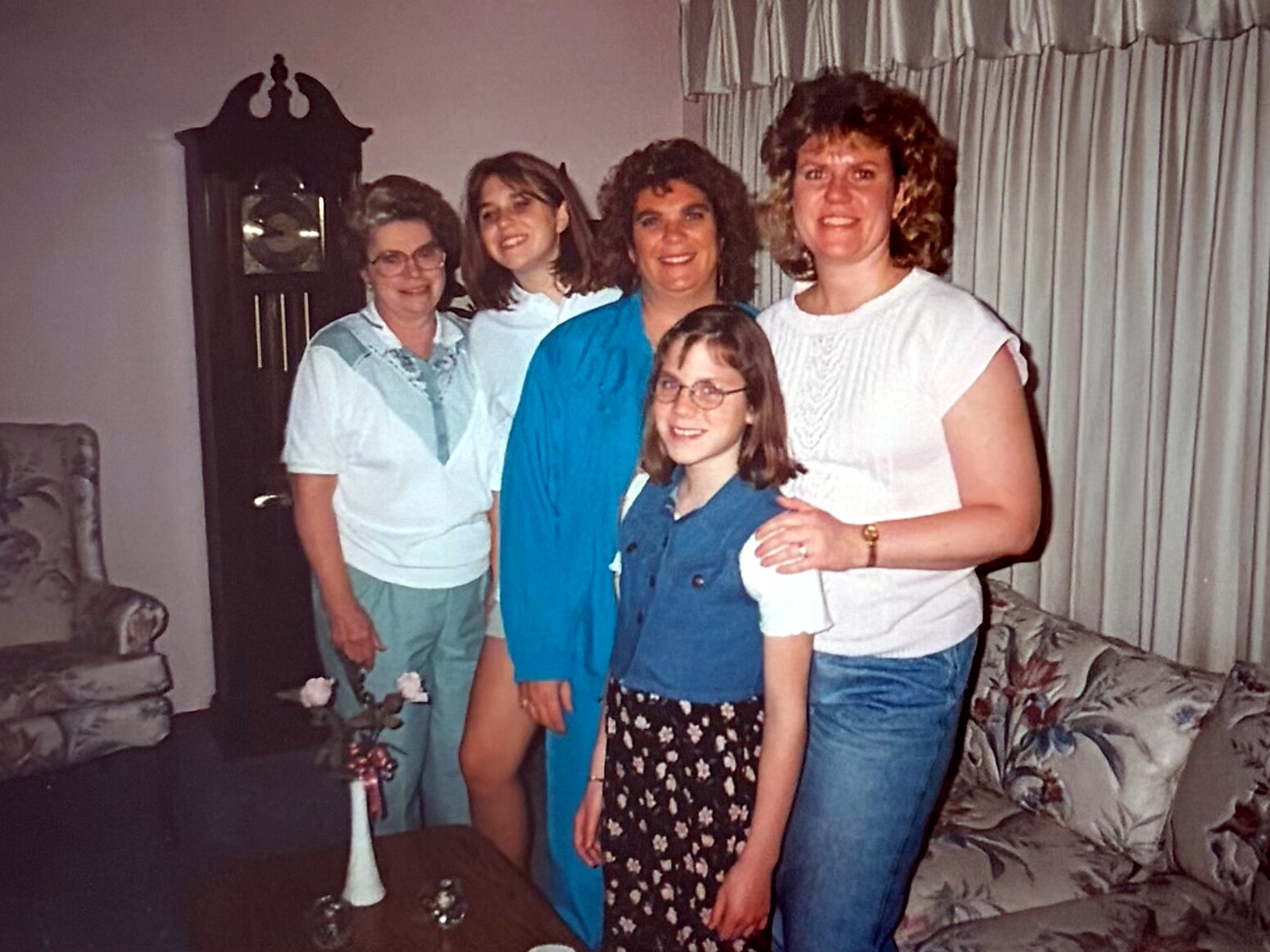 Five people posing together in a living room; a girl with glasses stands in front while four adults smile behind her, with a tall clock and curtains in the background.