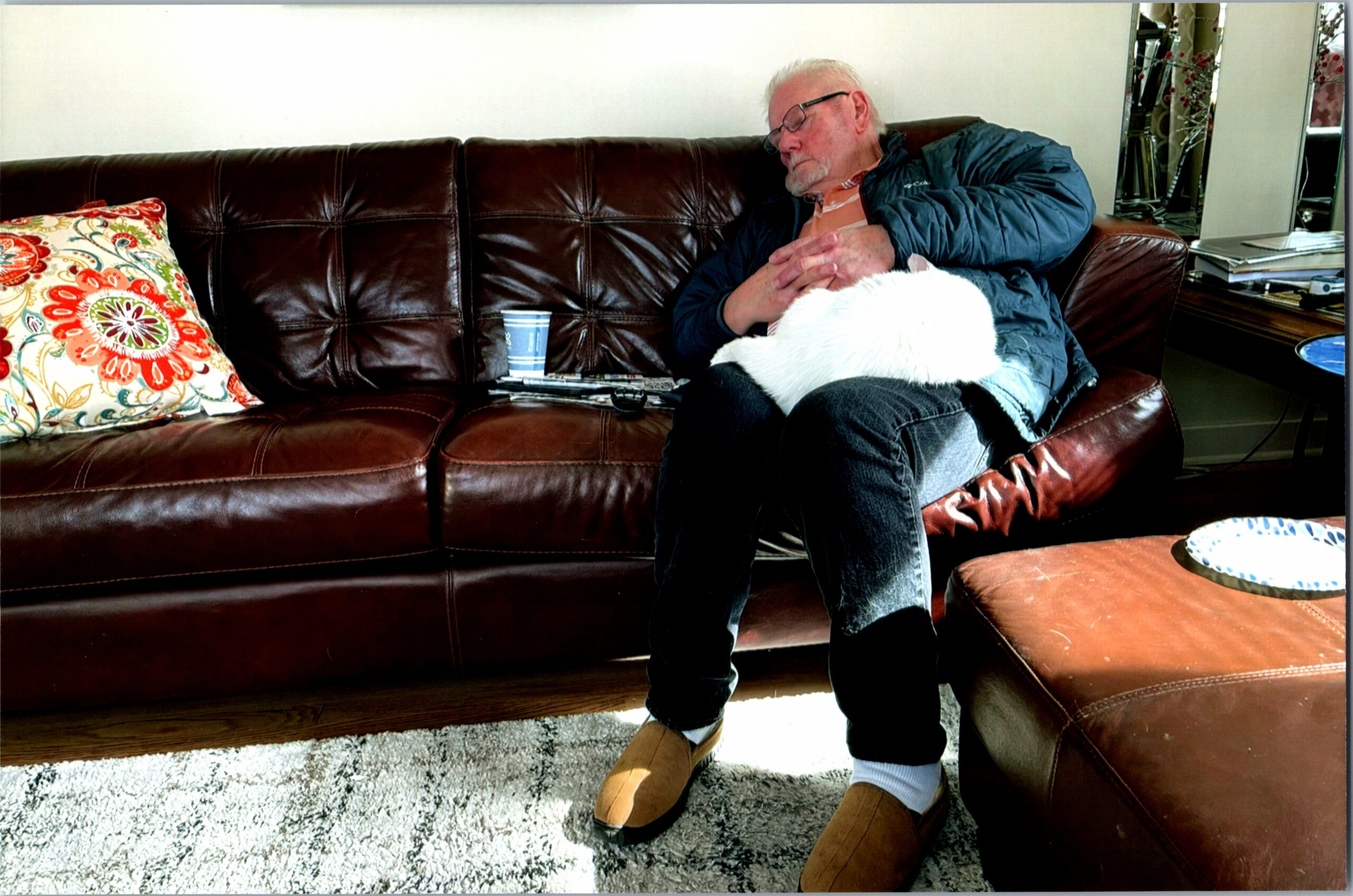 Older man with glasses sits on a brown leather couch, eyes closed, hands clasped on his chest in a living room setting.