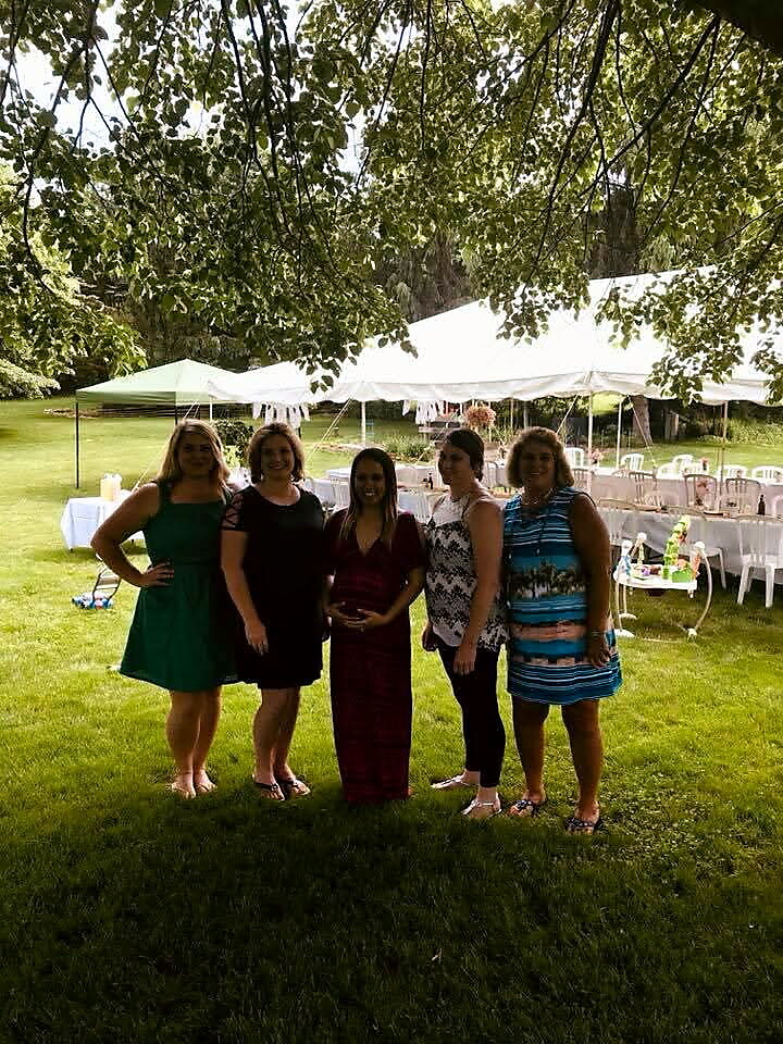 Five women pose together on a grassy lawn at an outdoor event with white tents in the background.