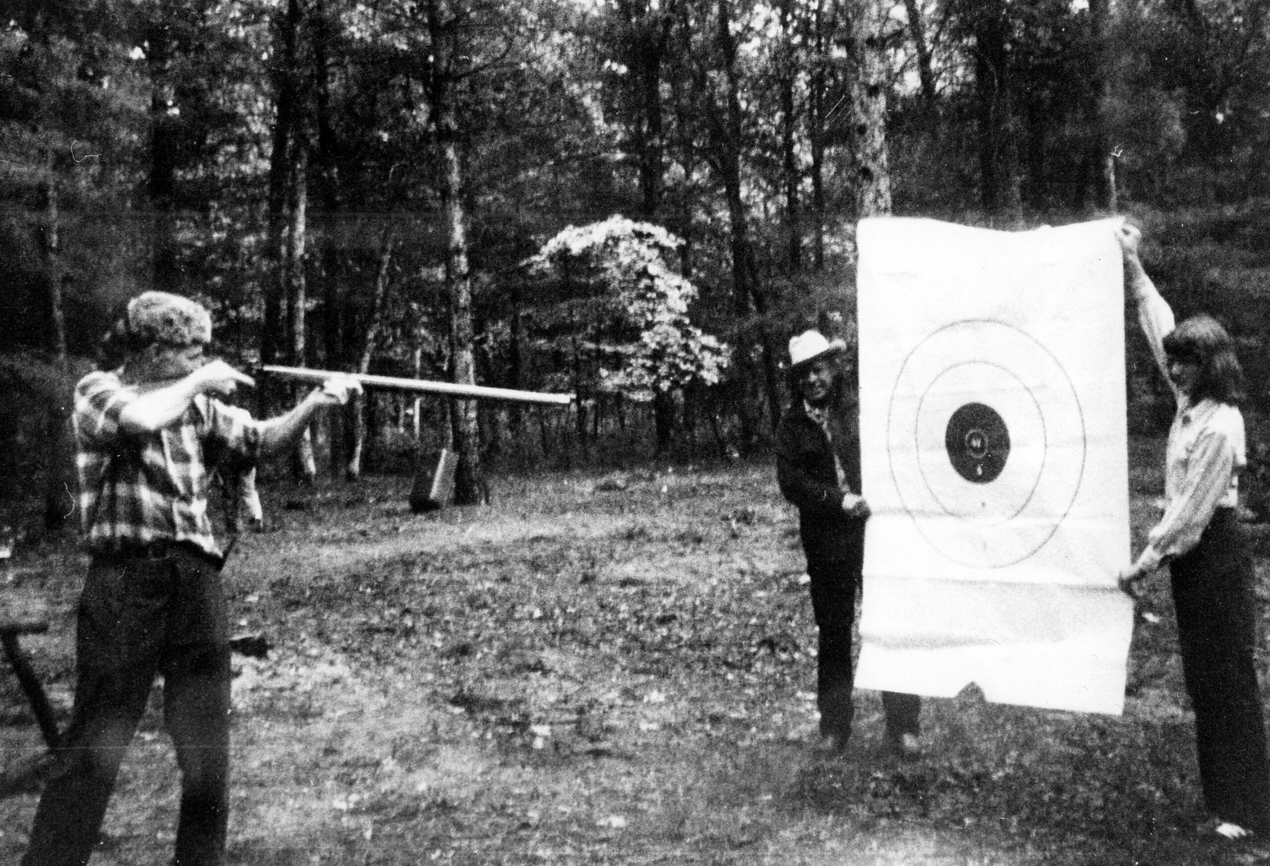A man with a rifle aims at a paper target in an outdoor wooded area, as bystanders watch nearby.