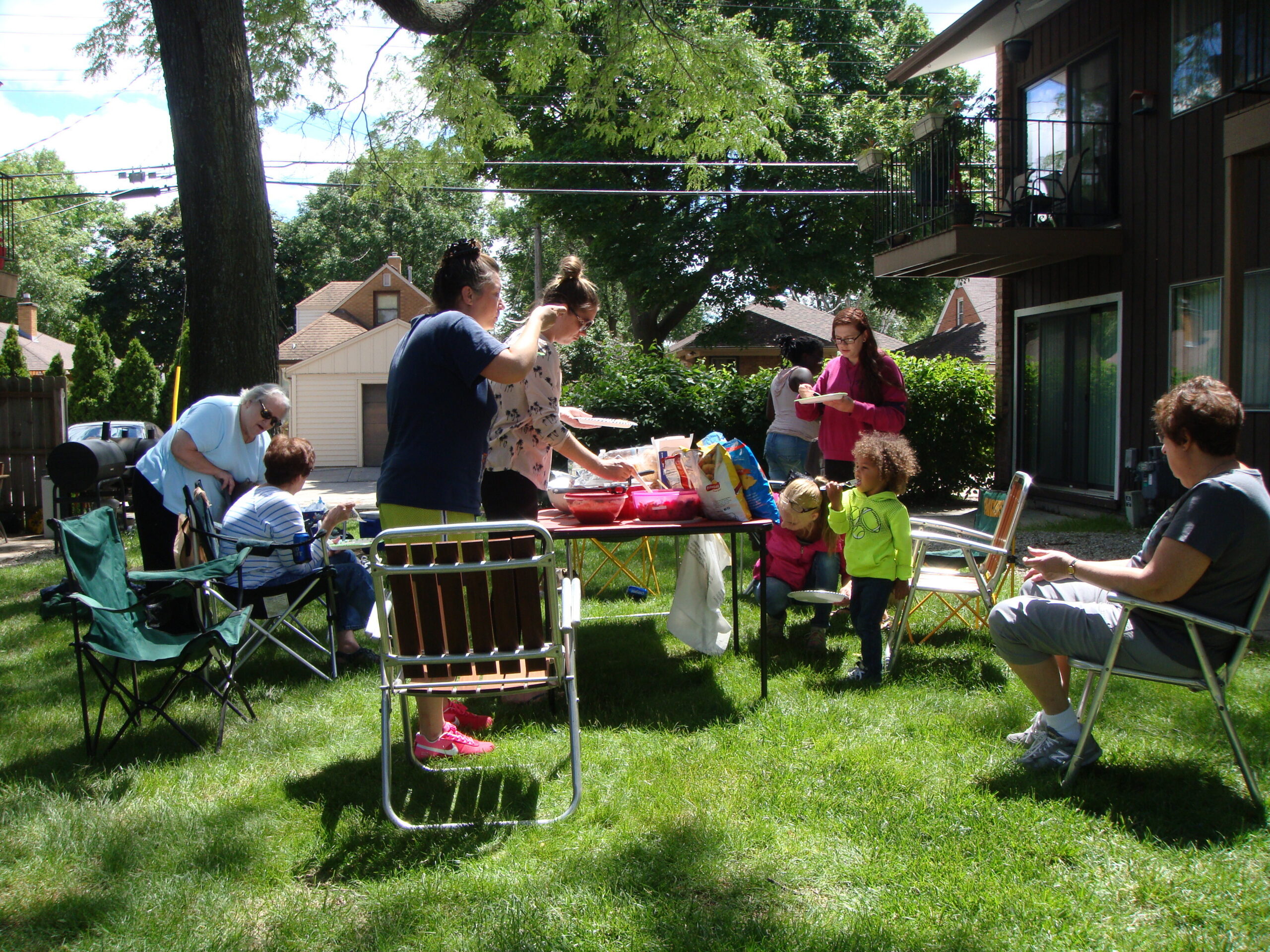Group of adults and children at a backyard cookout with folding chairs, a table of snacks, and sunny trees overhead.