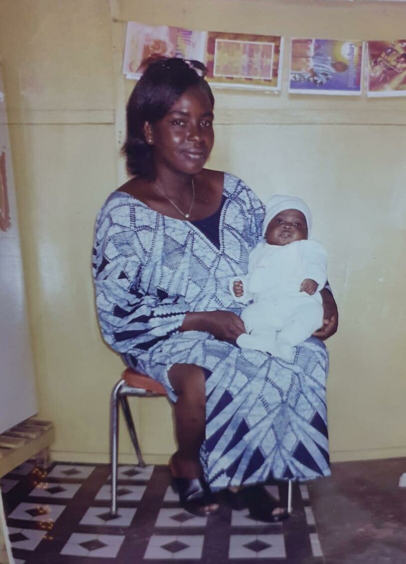 Woman in a blue and white patterned dress sits on a chair, cradling a baby in white clothing and a white hat inside a room with posters on the wall.