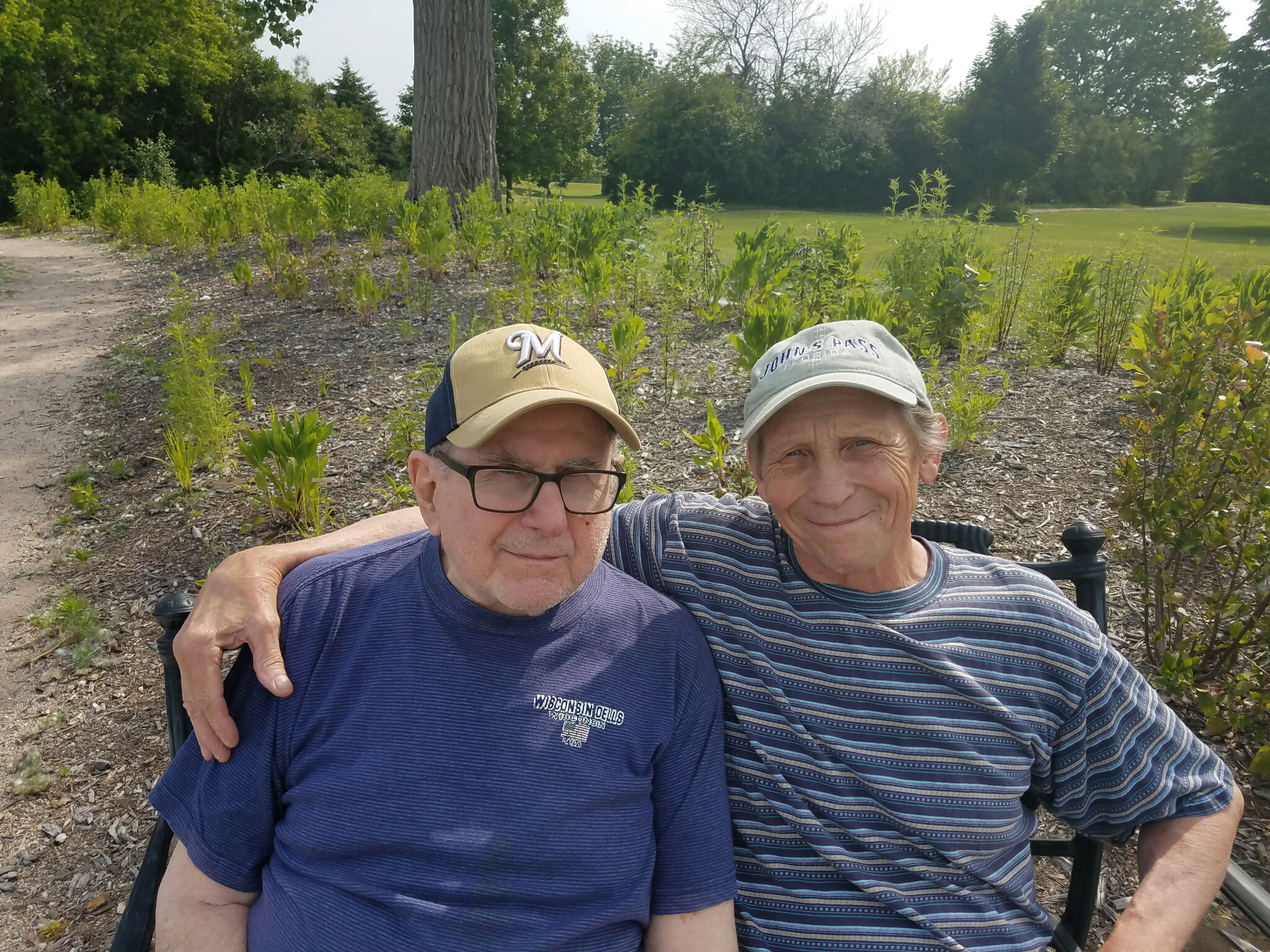 Two older men sit close together on a bench in a park, smiling at the camera.