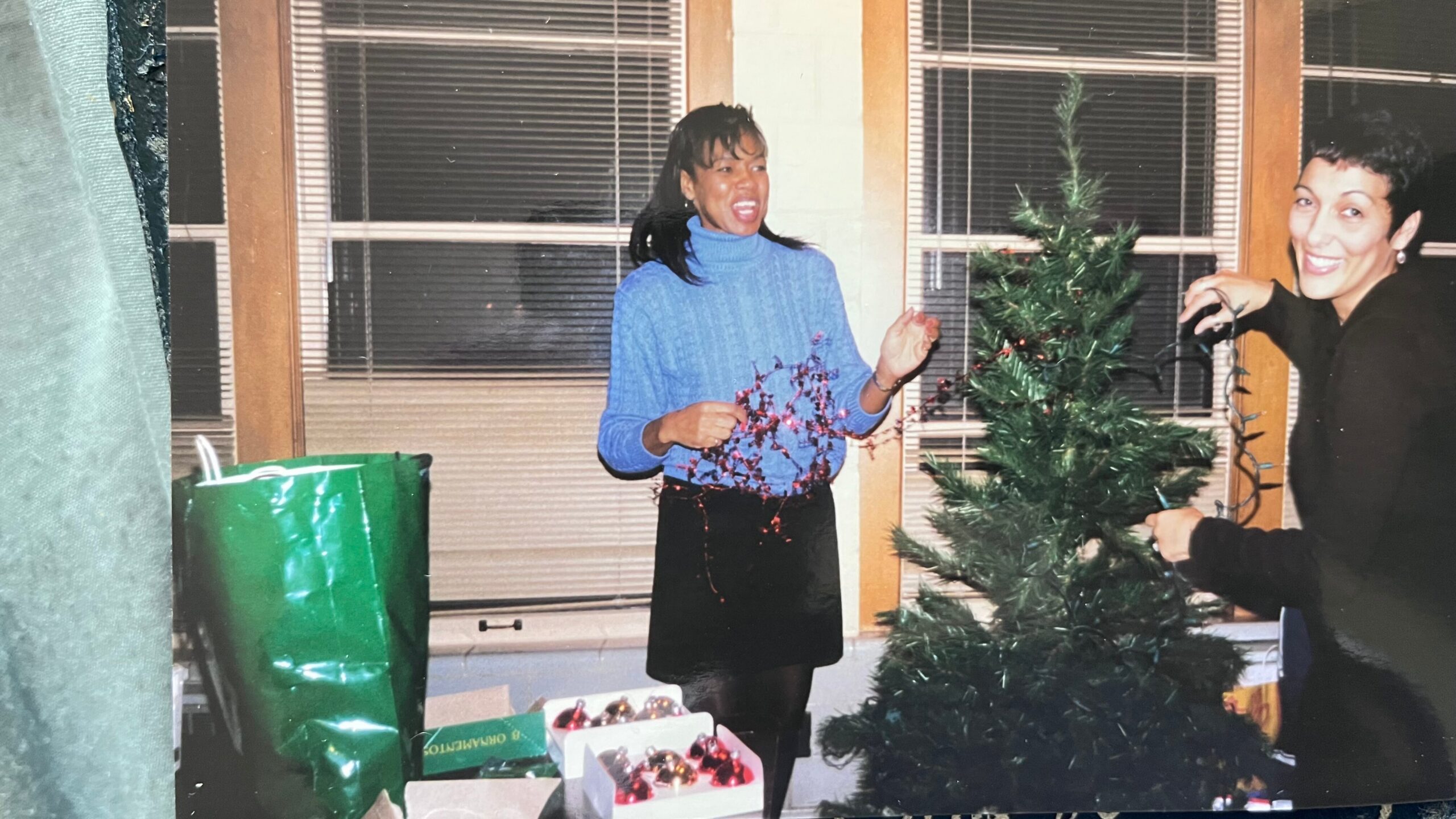 Two women decorating a Christmas tree indoors, smiling as they place garland and ornaments.