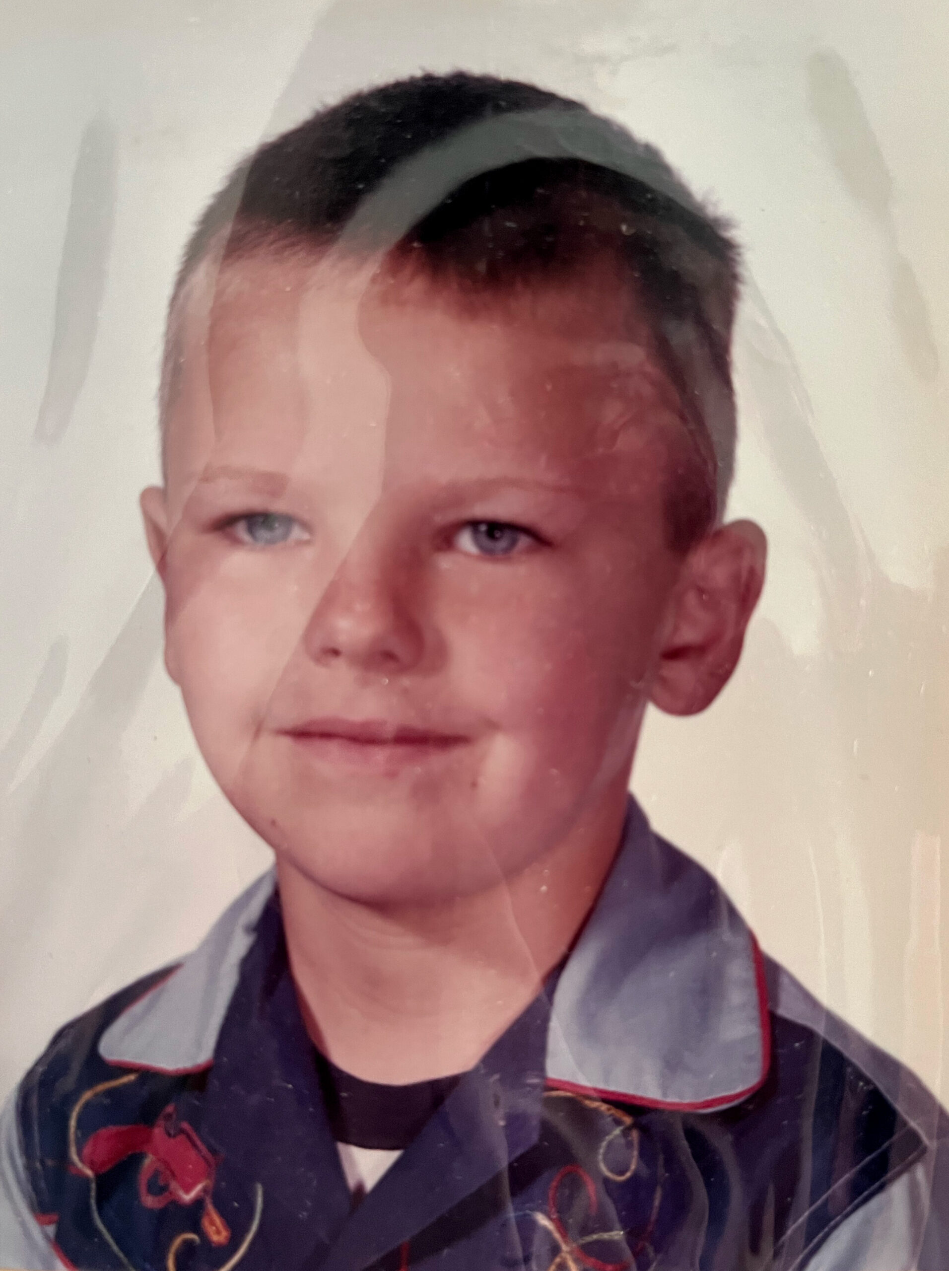 Portrait of a young boy with short dark hair, wearing a dark shirt with red and gold embroidery, looking at the camera (scan shows glare on photo).