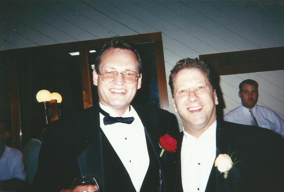 Two men in black tuxedos posing and smiling at a formal event, one with a red rose boutonniere and the other with a white rose boutonniere, with a guest in the background.