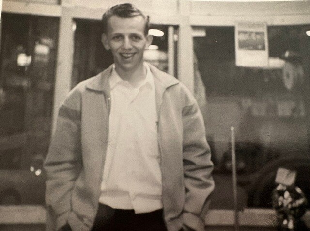 Smiling man in a light jacket and white shirt stands in front of a glass storefront.