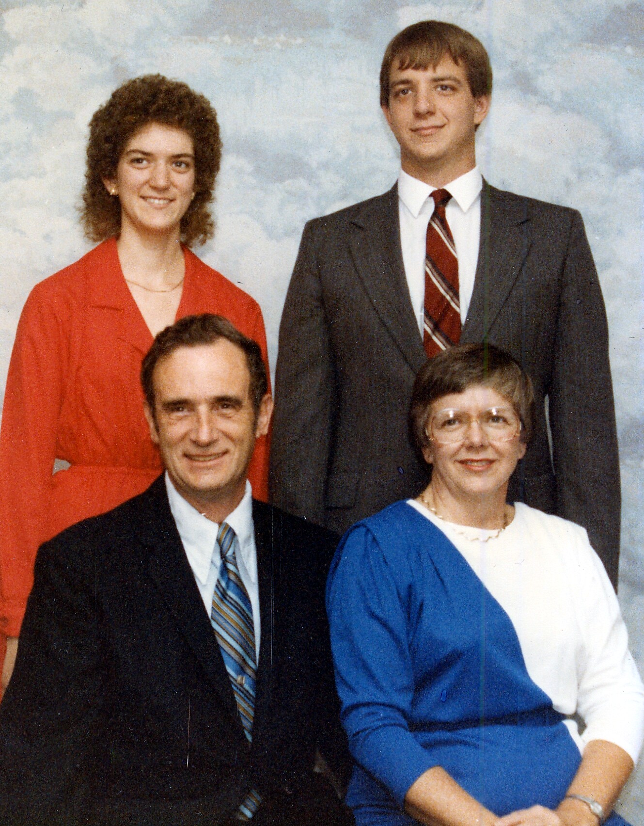Four adults posed for a formal group portrait: two seated in front (man in suit, woman in blue) and two standing behind (woman in red, man in suit)