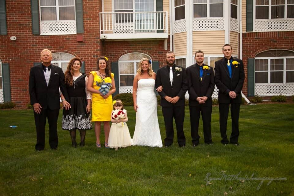 Wedding party posed in front of a brick building: bride in white gown center, groom and groomsmen in dark suits with yellow or blue boutonnieres, and a few family members on either side, standing on a green lawn.