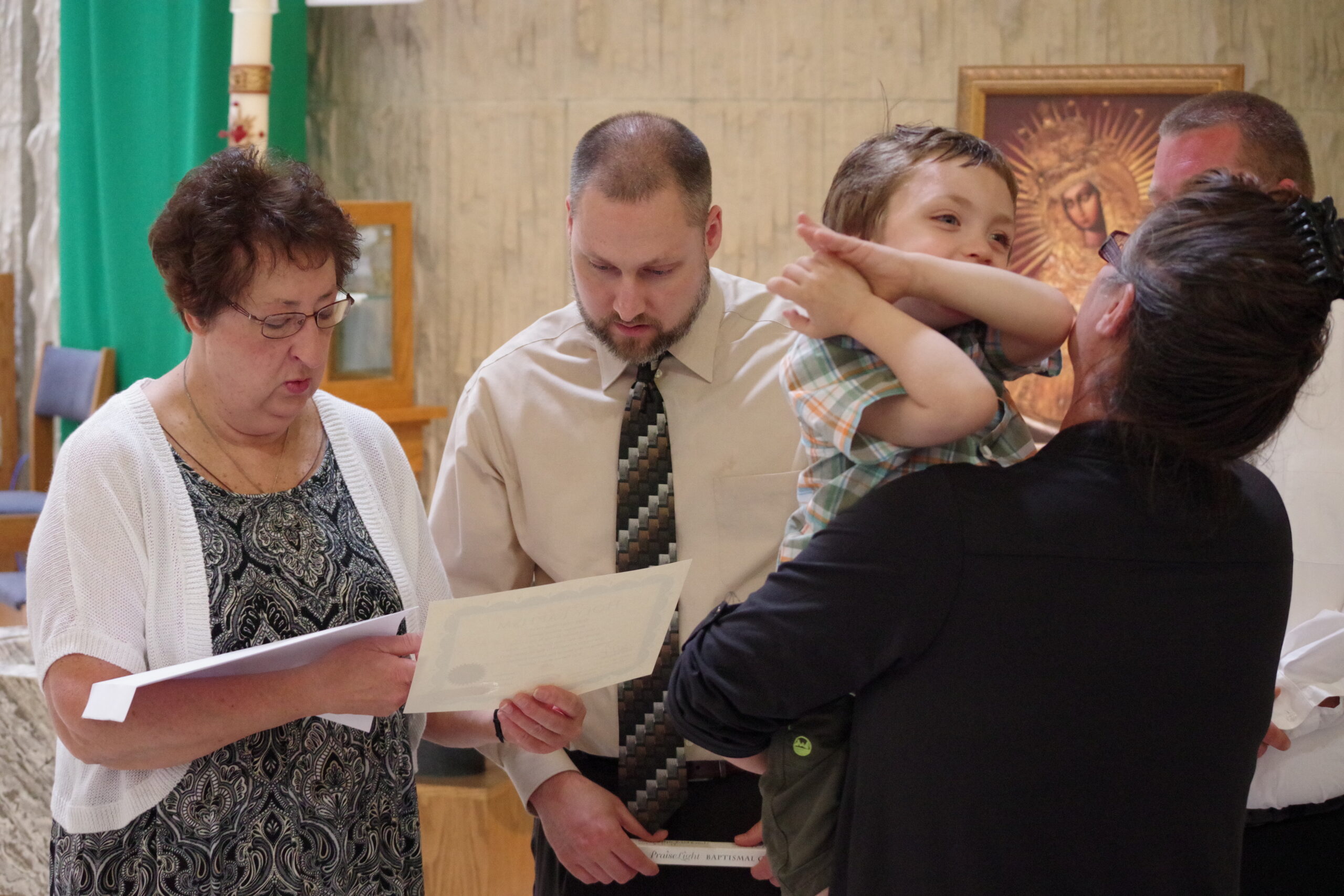 People gather around papers in a church-like setting; a woman in a white cardigan reads a document while a man in a shirt and tie looks on and a child is held nearby by another adult.
