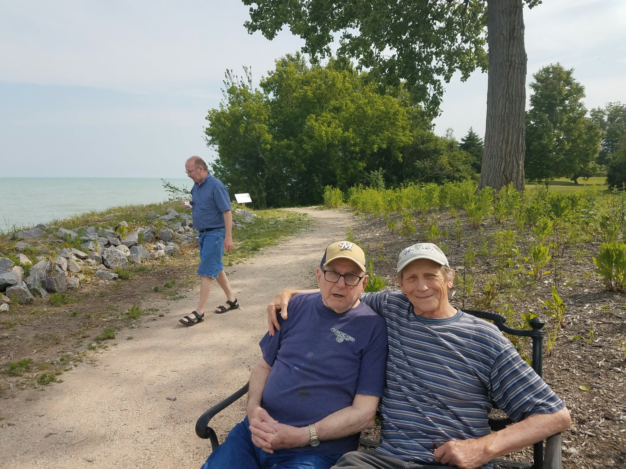 Two seniors in a wheelchair posing on a dirt path beside a lake, with a man walking and leafy trees in the background.”