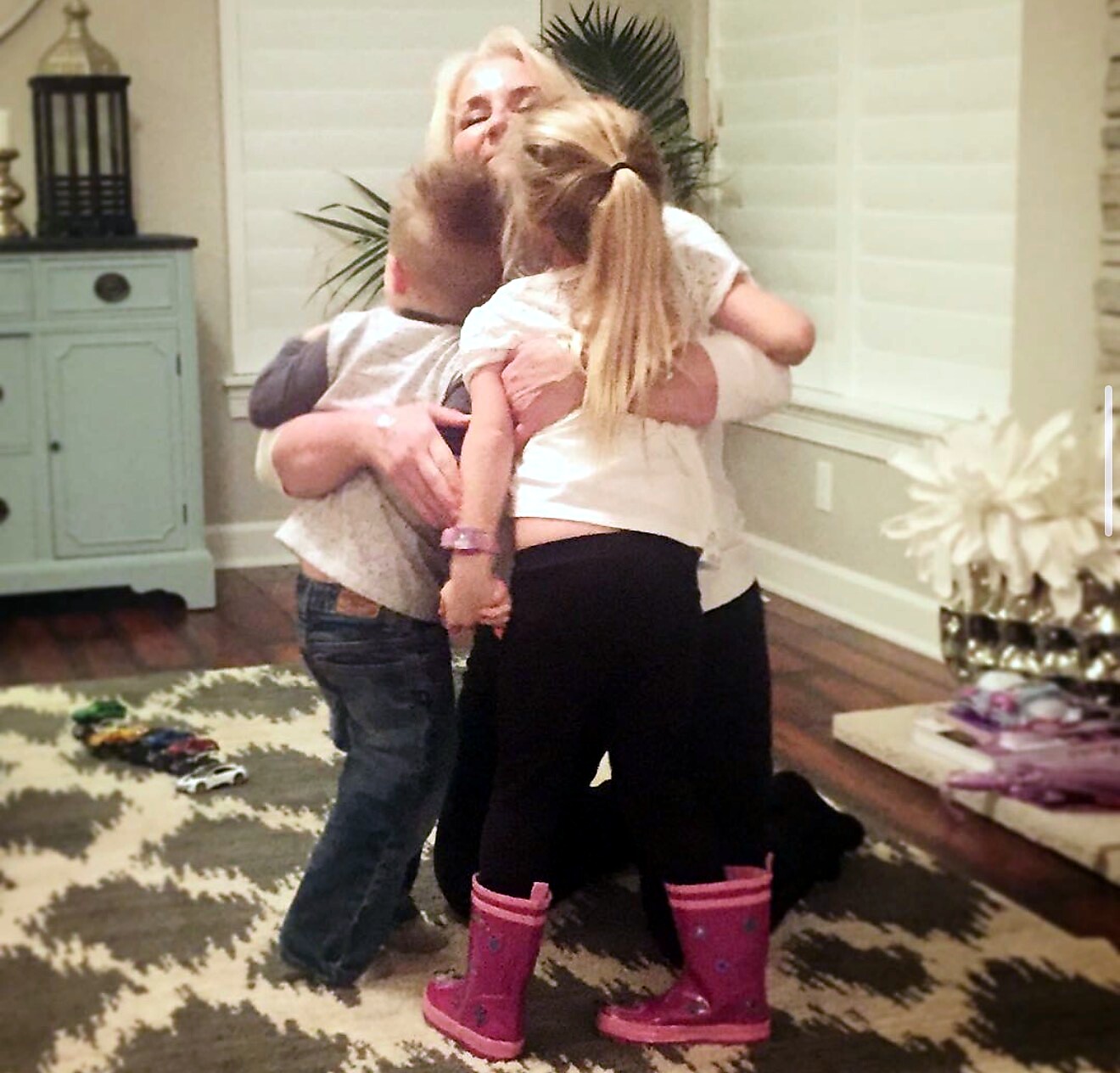 A family group hug in a cozy living room, with a smiling woman embracing two children.