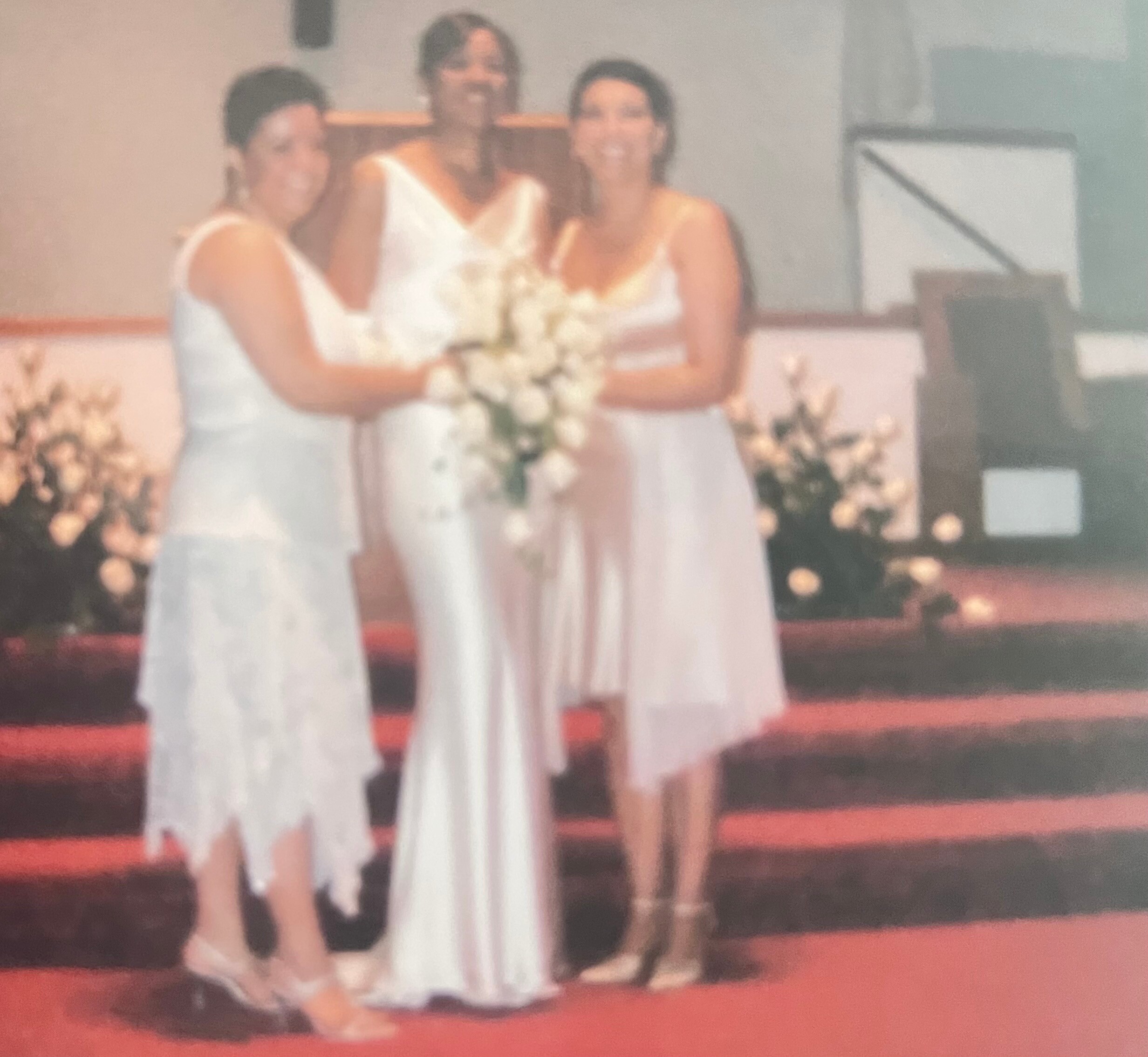 Two women in white dresses standing on a stage, smiling and holding bouquets during a ceremony or celebration.