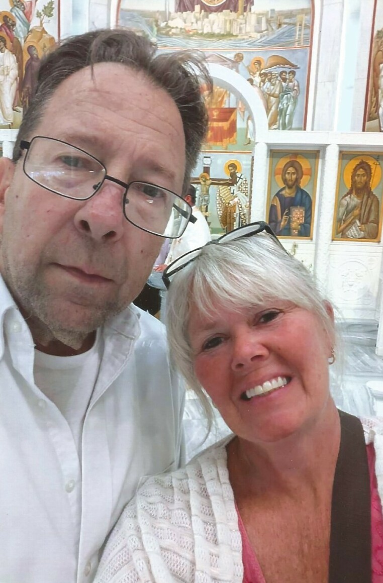 Close-up selfie of a man with glasses and a smiling woman in front of a wall of religious icons.