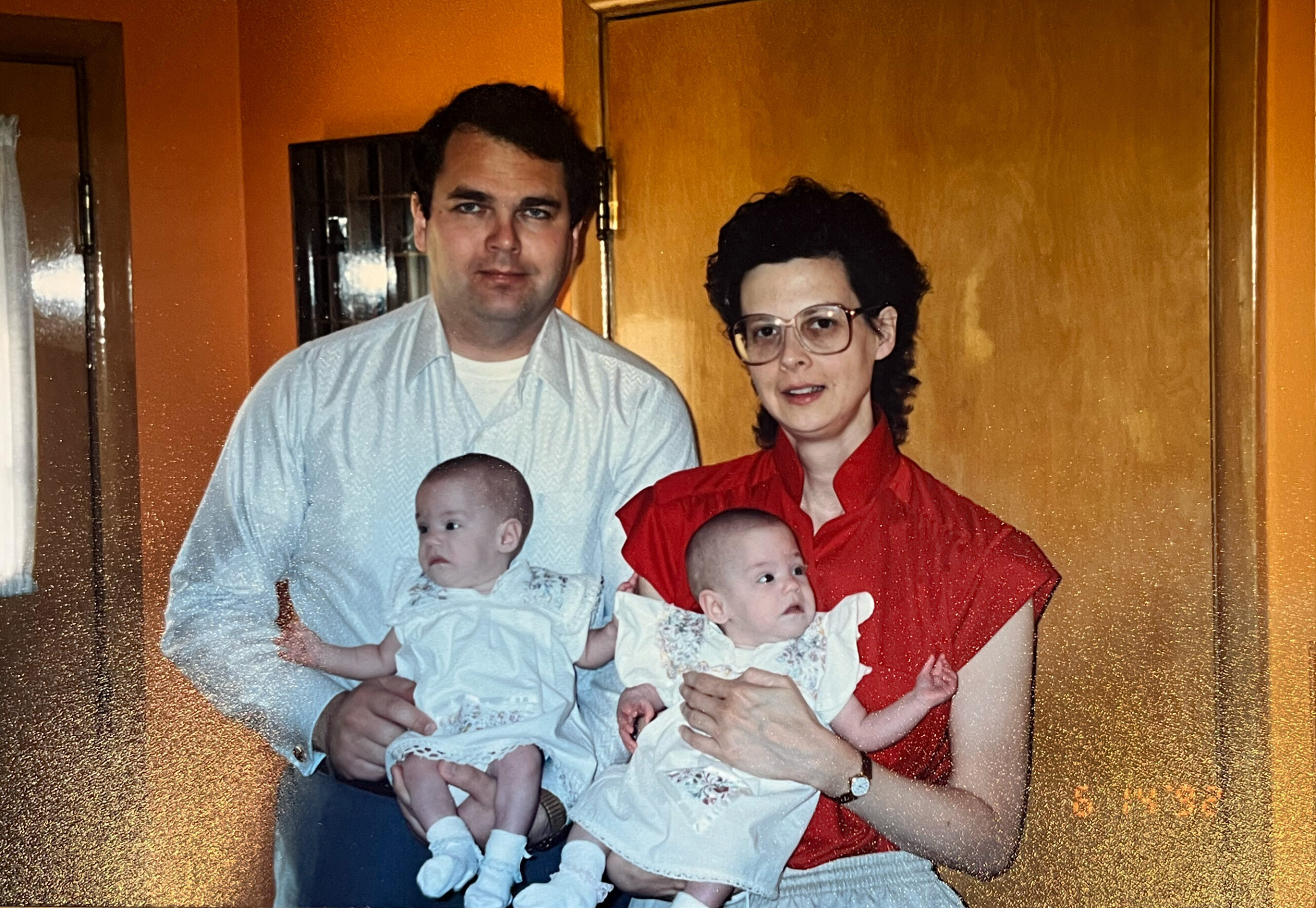 Father and mother hold two babies indoors, standing together in front of orange walls.