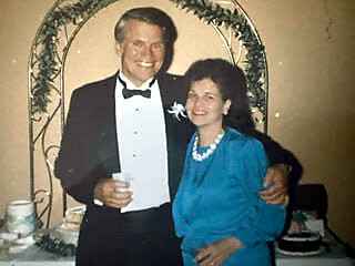 Smiling man in a tuxedo and a woman in a blue dress pose together at a wedding reception, near a floral arch.