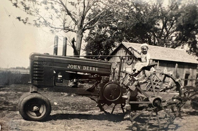 Vintage John Deere tractor with metal-spoked wheels in a farmyard, a person seated on the tractor near a wooden barn.