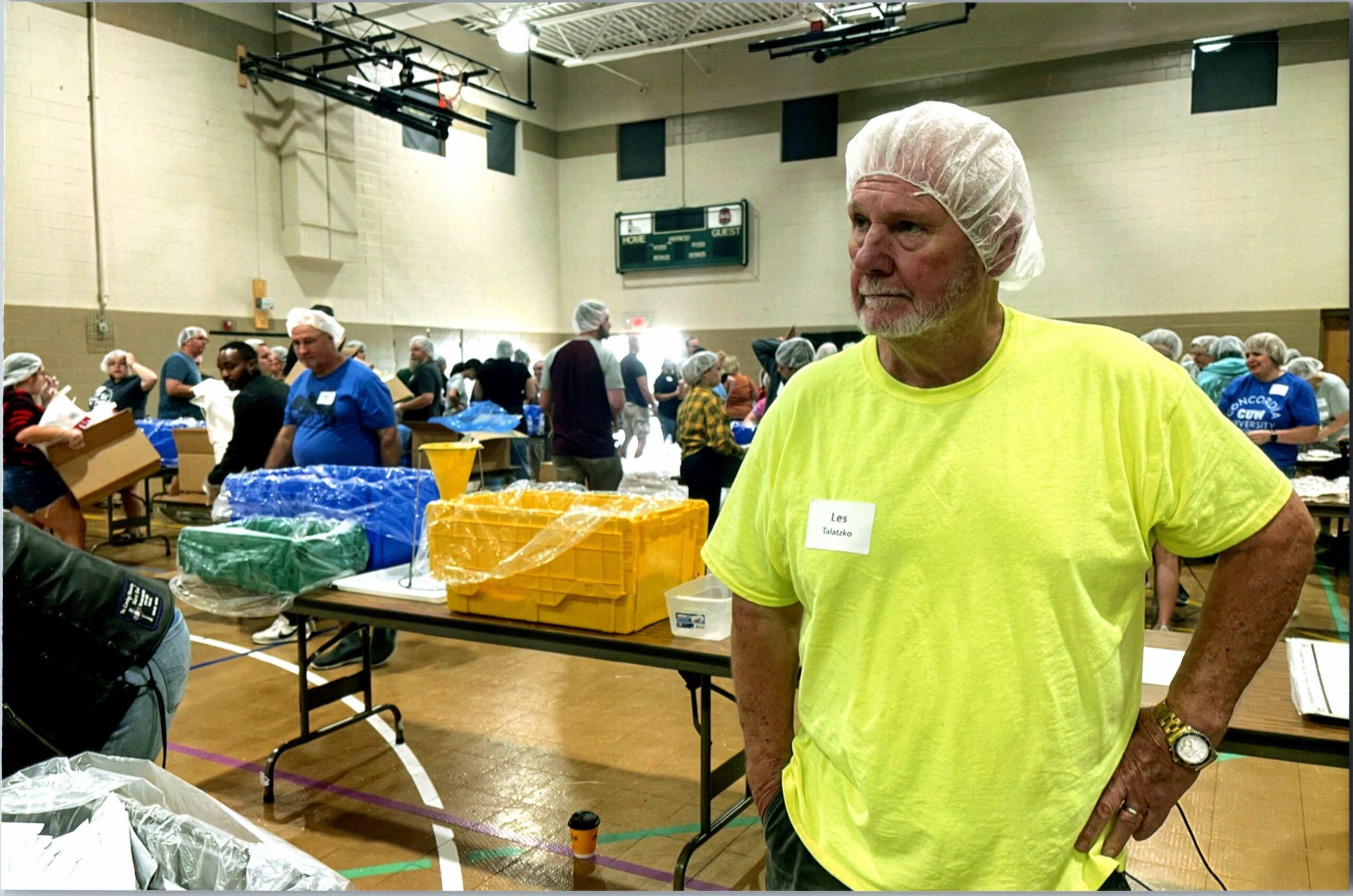 Man in a hairnet and neon yellow shirt stands at a table sorting supplies in a gym, with volunteers in the background.