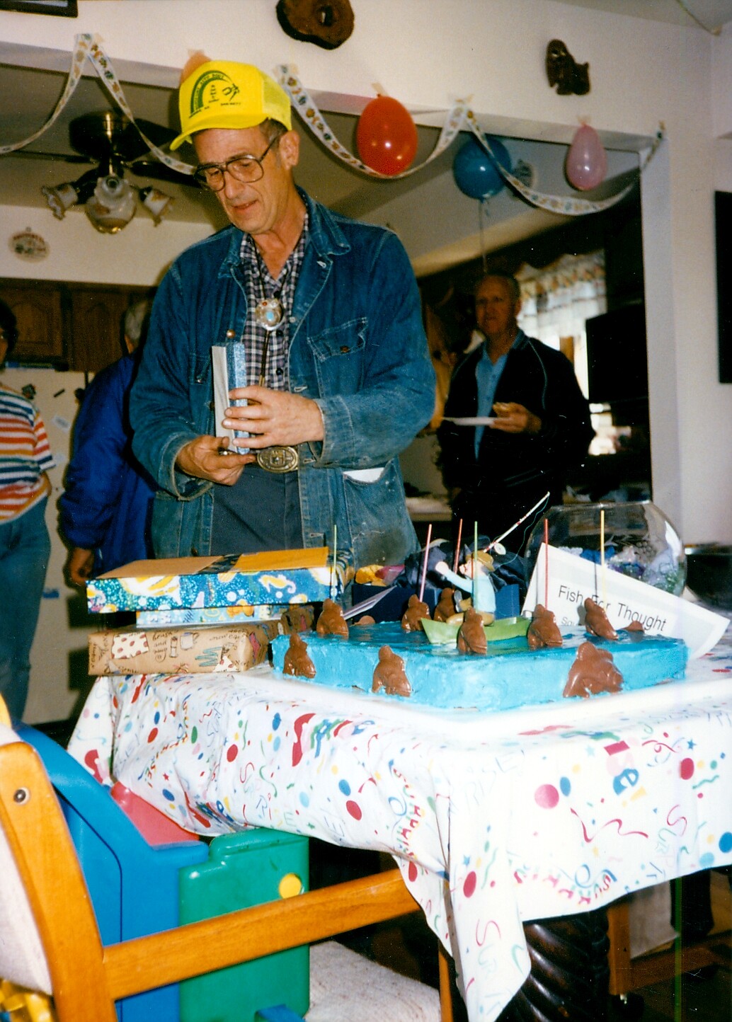 Man in a yellow birthday cap and denim jacket cuts a blue cake topped with small chocolate figures at a party, with gifts and decorations in the background.