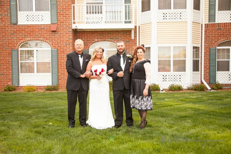 Bride in a white wedding gown holds a red-rose bouquet, standing with her groom and two other adults in formal wear on a grassy lawn in front of a brick building.