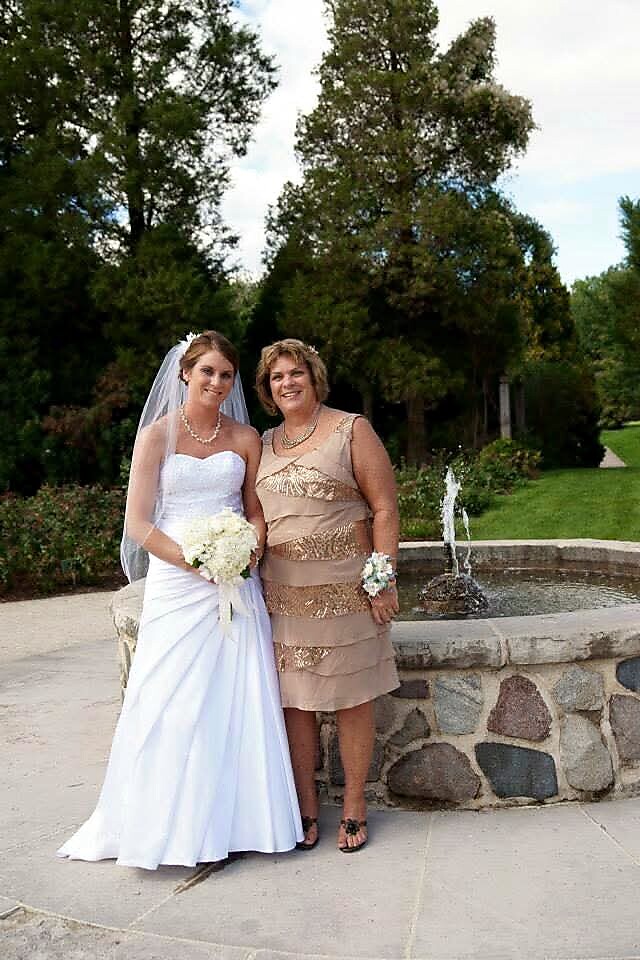 Bride in a white wedding gown with bouquet stands beside a guest in a beige dress outdoors by a stone fountain.