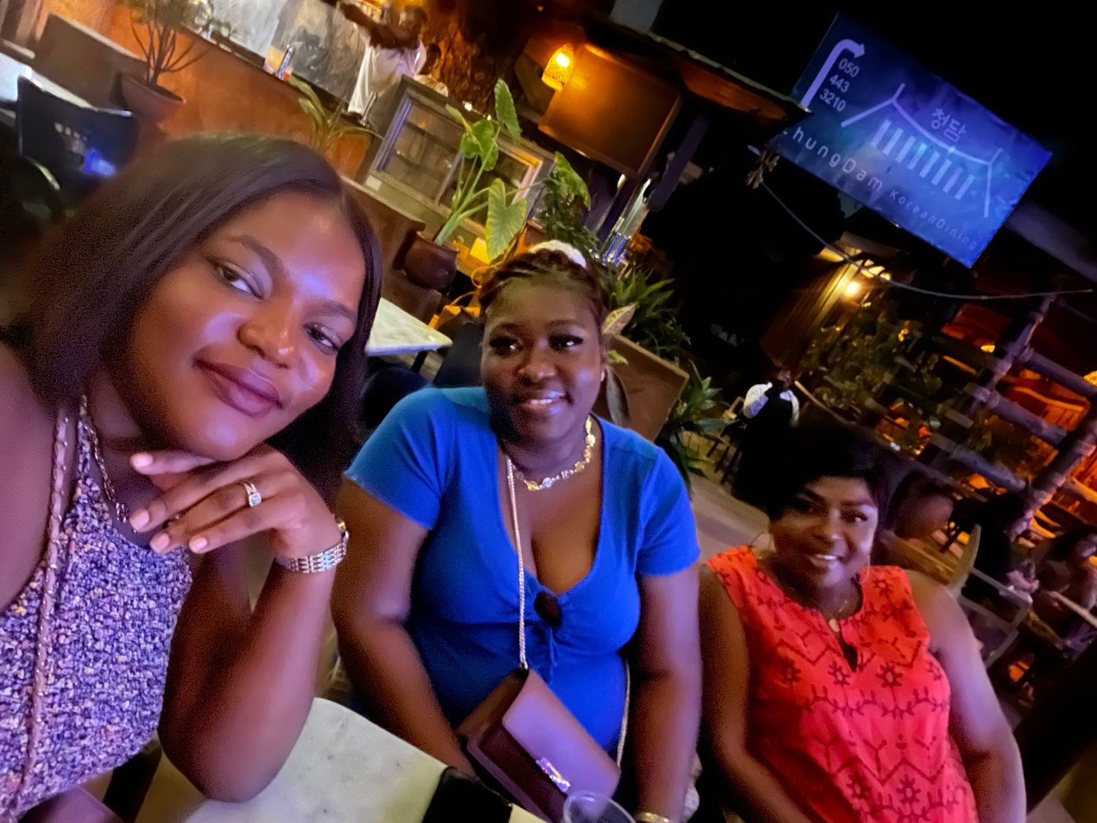 Three women sit together at an outdoor night restaurant, smiling at the camera with drinks on the table.