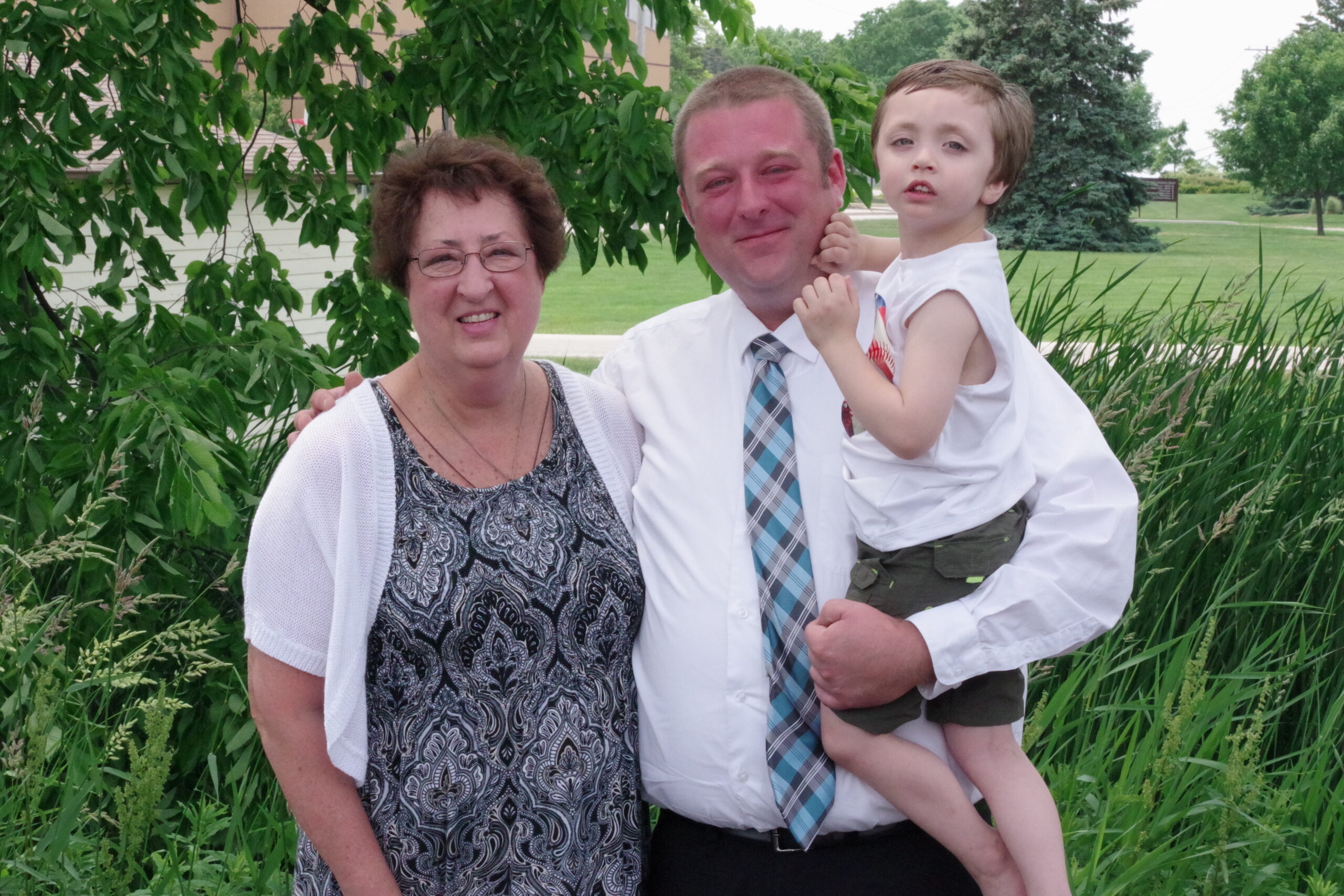 Family of three posing outdoors: grandmother on the left, father in the middle, and his young son being carried, with greenery behind them.