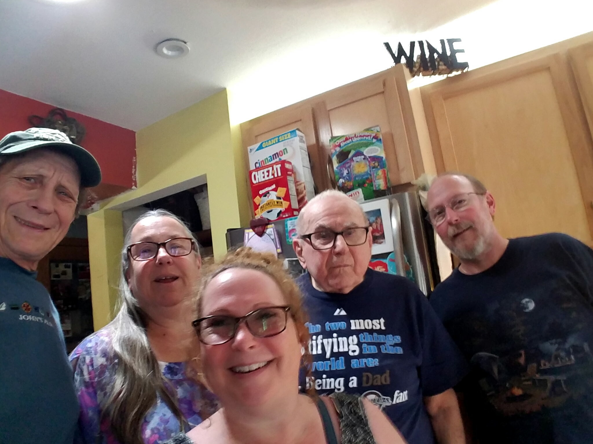 Group of adults taking a casual kitchen selfie, smiling at the camera, with cereal boxes and a 'WINE' sign on the cabinets in the background.