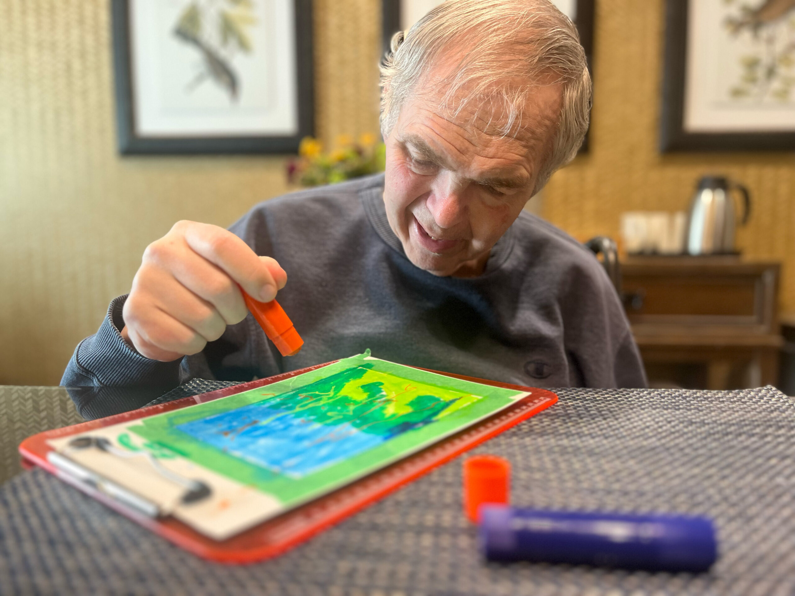 Older man with gray hair concentrating on a bright, colorful drawing using an orange marker at a table indoors.