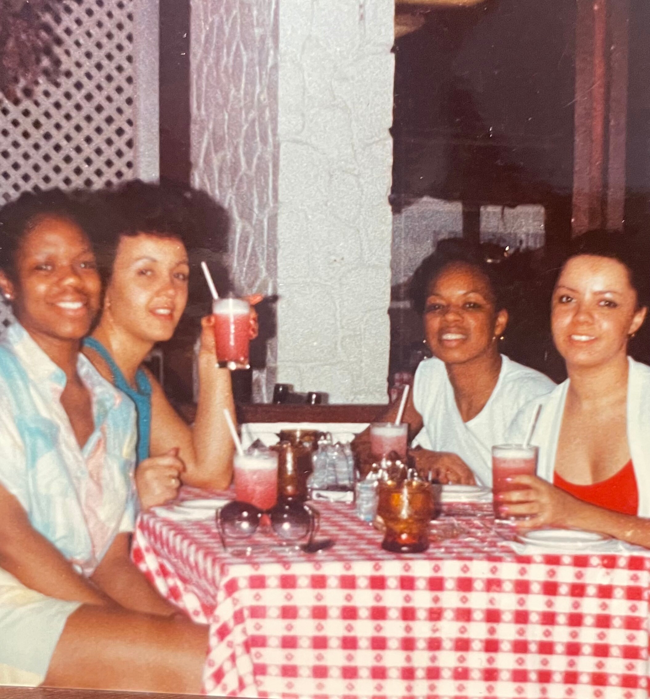 Four friends sit around a table with a red-and-white checkered tablecloth, smiling at the camera with drinks in hand.