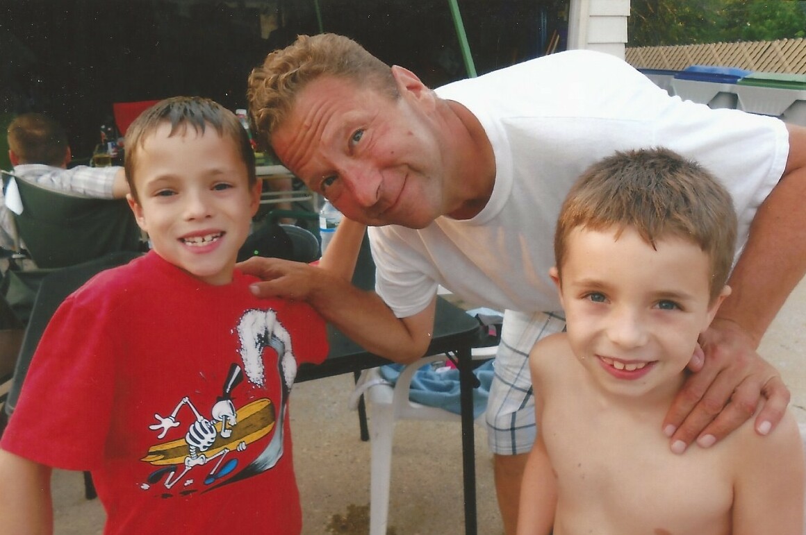 Man bending down with two smiling boys at a backyard party.