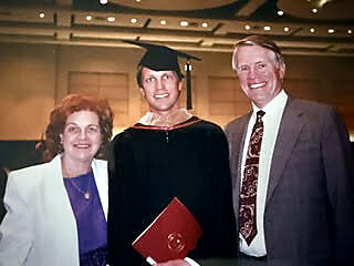 Graduate in cap and gown posing with two smiling adults at a ceremony, one holding a red diploma cover in front of a warmly lit crowd.