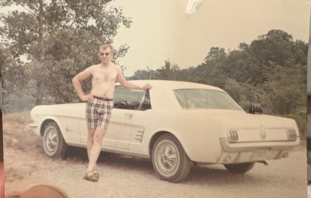 Shirtless man in plaid shorts leaning on a white vintage convertible on a dirt road, trees in the background.