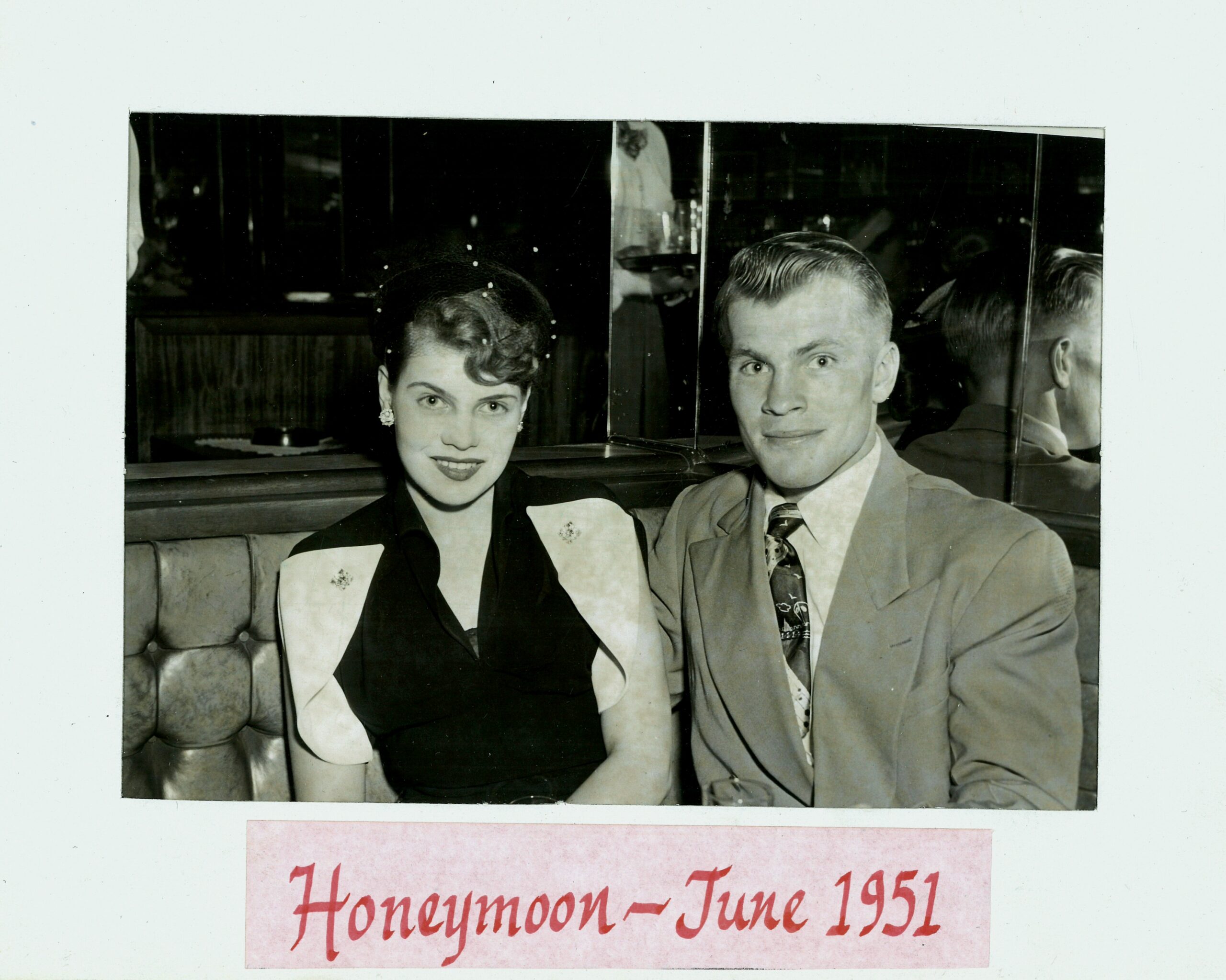Black-and-white photo of a smiling couple in a car, woman in vintage dress and pearls, man in suit; label reads 