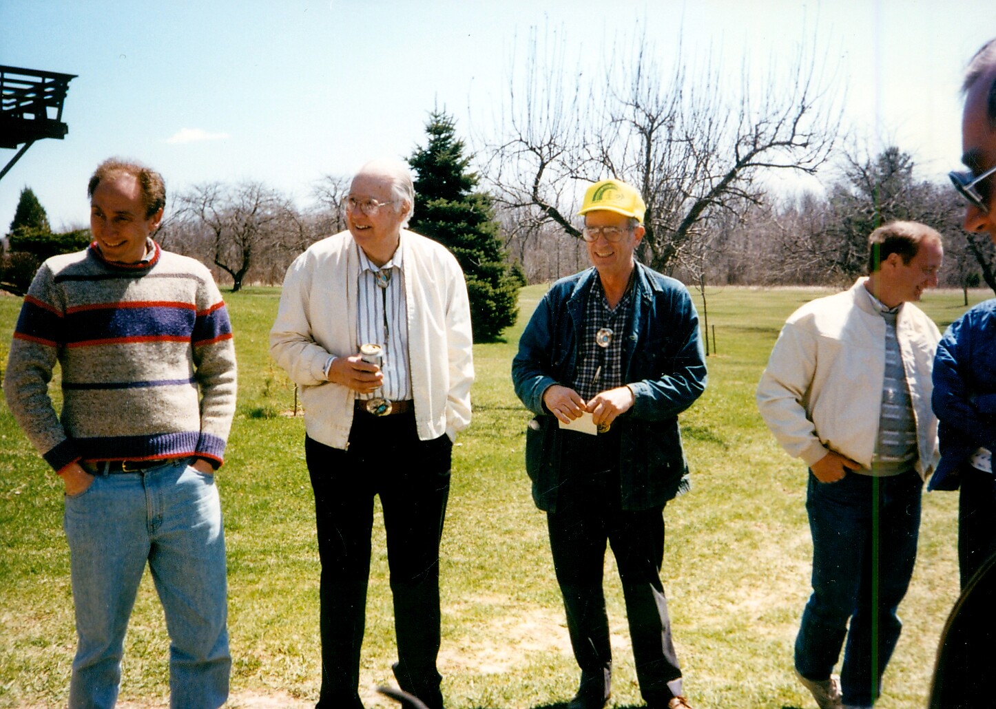 Group of men standing on a sunny lawn, casually talking and laughing together in a park-like setting.