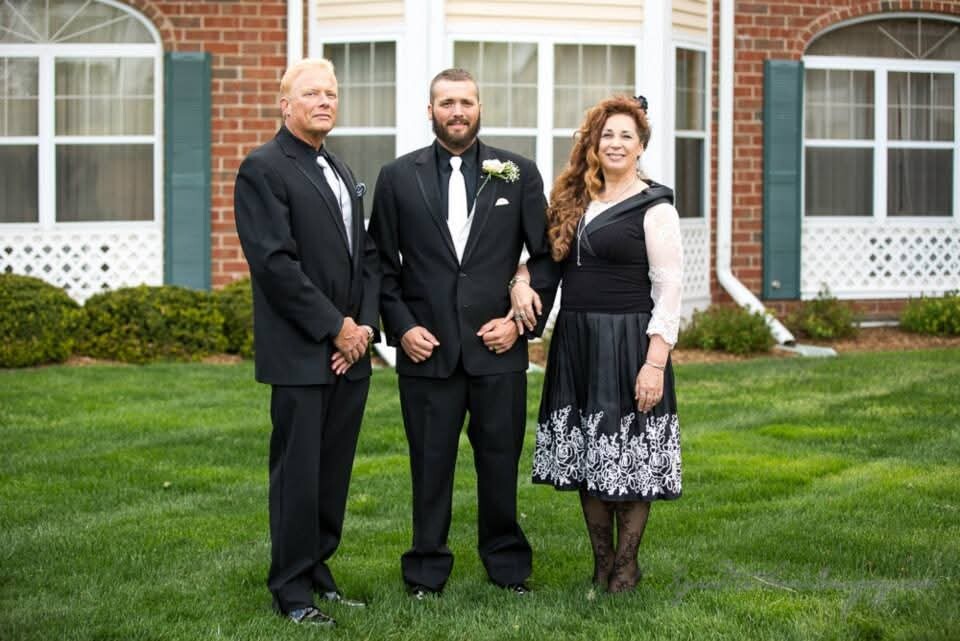 Three adults in formal attire pose on a green lawn in front of a brick building; center man has boutonniere, others in suits and a dress outfit behind him.