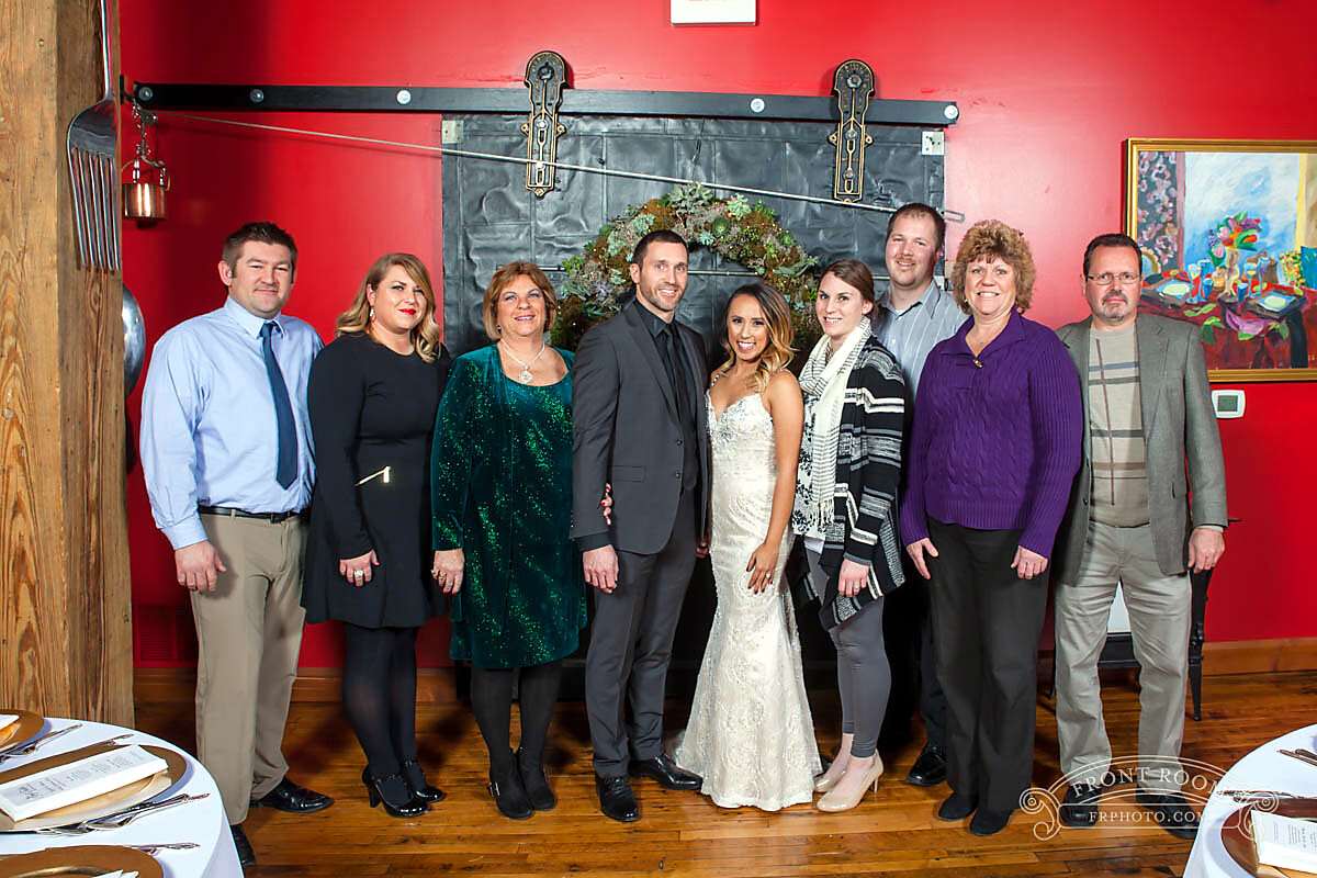 Wedding party group photo: bride and groom centered, surrounded by family and friends indoors in a red-walled room with a wreath backdrop behind them.